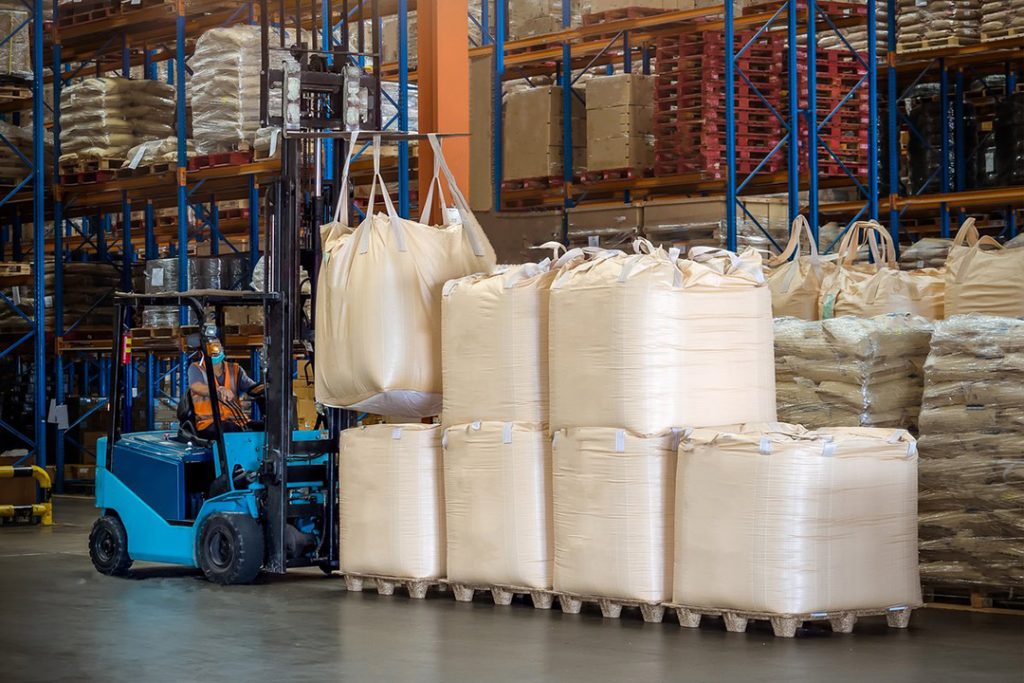 A blue forklift picking up a loaded bulk bag from a large stack inside a warehouse