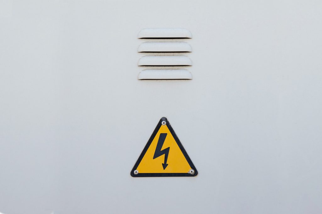 Grey metal door with louvres and a yellow electric warning sign.