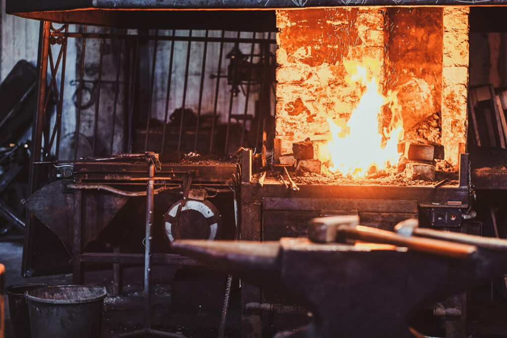 Inside a blacksmith's tavern with a fire burning in the background and a workbench with a hammer and anvil in the foreground.