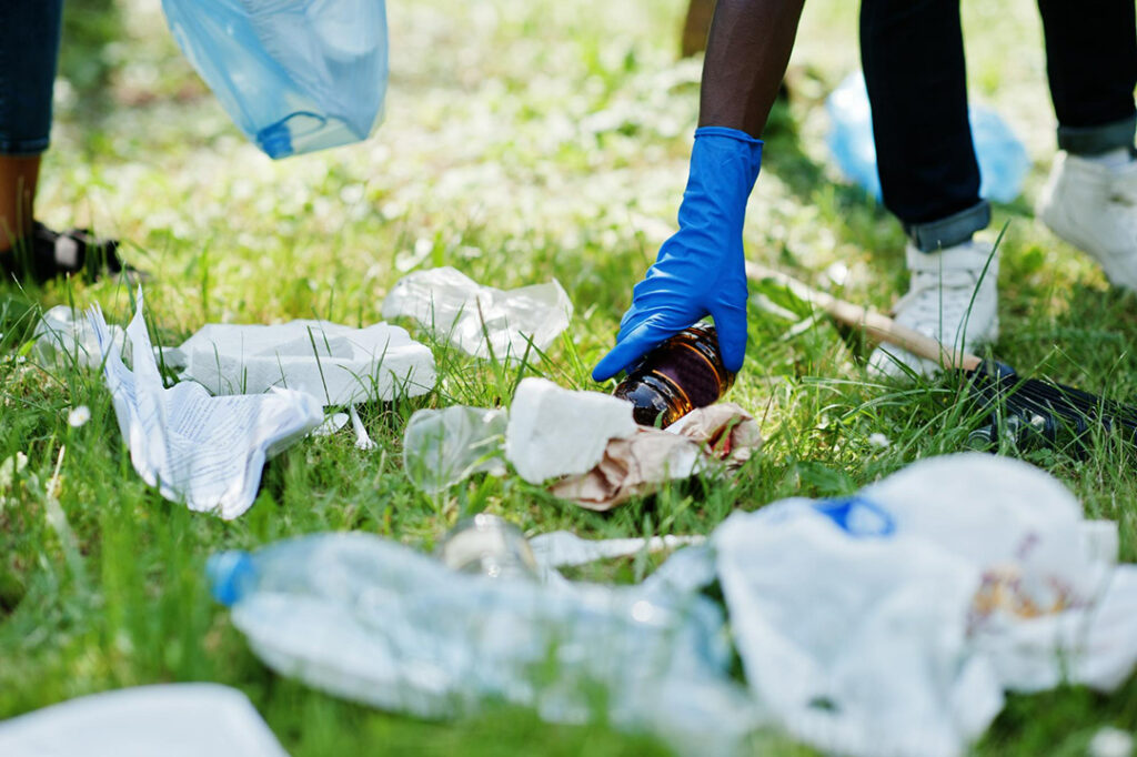 A man wearing blue rubber gloves picks up a glass bottle in an outdoor setting.